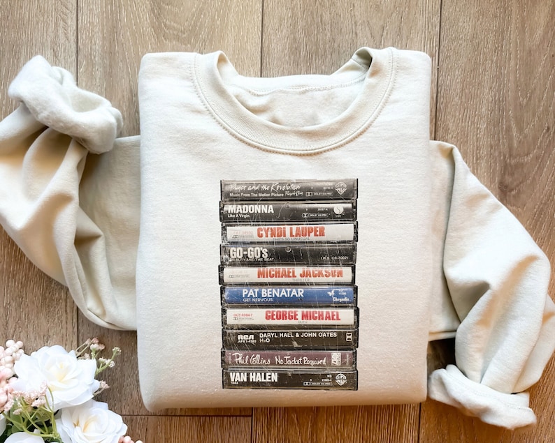 May include: A light beige sweatshirt with a stack of vintage cassette tapes printed on the front. The tapes feature artists such as Madonna, Cyndi Lauper, Go-Gos, Michael Jackson, Pat Benatar, George Michael, Daryl Hall & John Oates, Phil Collins, and Van Halen.