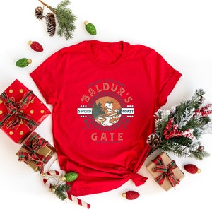 Op de afbeelding: Rood T-shirt met een witte grafische print van een cirkel met de tekst "Baldur's Gate" en een berglandschap in de cirkel. De tekst "Sword Coast" staat boven de cirkel en de tekst "Sword Coast" staat onder de cirkel. Het T-shirt is omringd door kerstversiering, waaronder dennentakken, rode en gouden cadeaudozen en snoepstokken.