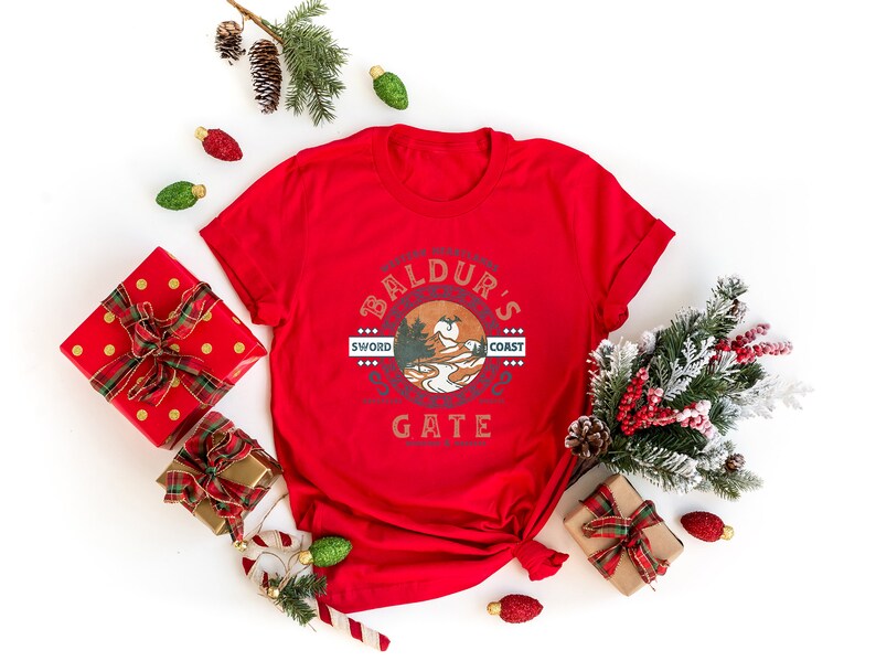 Op de afbeelding: Rood T-shirt met een witte grafische print van een cirkel met de tekst "Baldur's Gate" en een berglandschap in de cirkel. De tekst "Sword Coast" staat boven de cirkel en de tekst "Sword Coast" staat onder de cirkel. Het T-shirt is omringd door kerstversiering, waaronder dennentakken, rode en gouden cadeaudozen en snoepstokken.