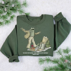 Peut inclure: Sweat-shirt vert avec un motif de deux grenouilles. Le texte sur le sweat-shirt est "WE MUST STOP EATING!" "CRIED TOAD AS HE ATE ANOTHER." - ARNOLD LOBEL