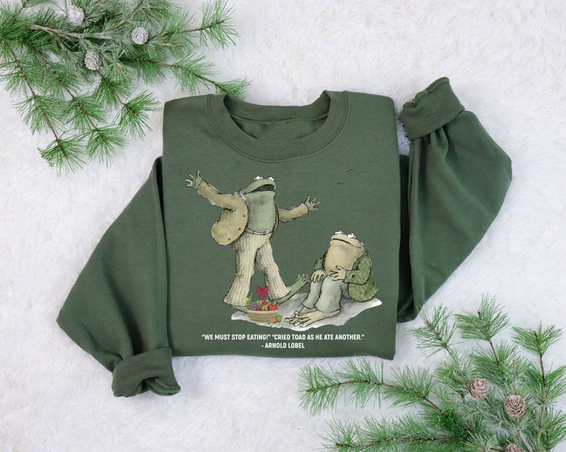 Peut inclure: Sweat-shirt vert avec un motif de deux grenouilles. Le texte sur le sweat-shirt est "WE MUST STOP EATING!" "CRIED TOAD AS HE ATE ANOTHER." - ARNOLD LOBEL