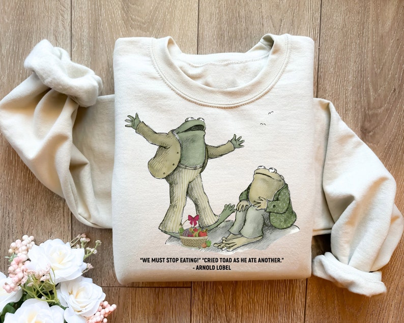 Peut inclure: Un sweat-shirt beige avec une illustration de grenouille verte et la citation "WE MUST STOP EATING!" "CRIED TOAD AS HE ATE ANOTHER." - ARNOLD LOBEL