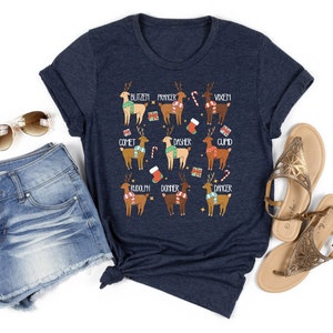 May include: Navy blue t-shirt featuring a festive design with cartoon reindeer, each labeled with a name like Blitzen, Prancer, and Vixen. The reindeer are adorned with scarves and surrounded by holiday-themed graphics. The shirt is paired with denim shorts and sandals.