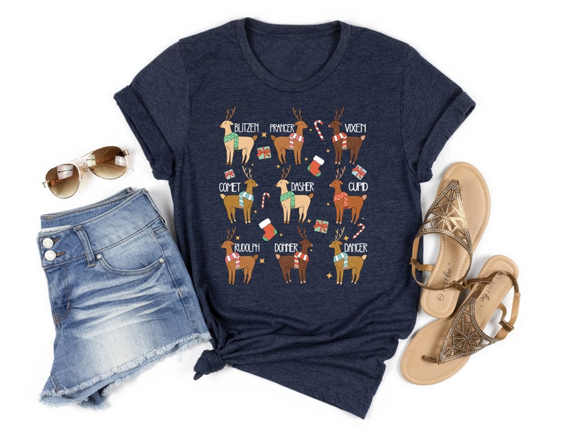 May include: Navy blue t-shirt featuring a festive design with cartoon reindeer, each labeled with a name like Blitzen, Prancer, and Vixen. The reindeer are adorned with scarves and surrounded by holiday-themed graphics. The shirt is paired with denim shorts and sandals.