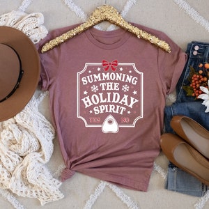 May include: A mauve t-shirt with the text "SUMMONING THE HOLIDAY SPIRIT" in a white, decorative frame. The shirt is styled with a brown hat, cream-colored scarf, blue jeans, and brown flats. A gold sequined hanger holds the shirt.