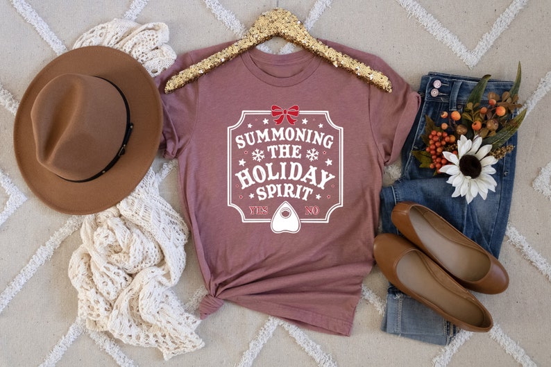 May include: A mauve t-shirt with the text "SUMMONING THE HOLIDAY SPIRIT" in a white, decorative frame. The shirt is styled with a brown hat, cream-colored scarf, blue jeans, and brown flats. A gold sequined hanger holds the shirt.