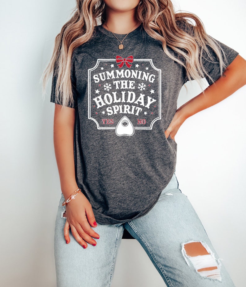 May include: Dark gray t-shirt with a white and red graphic. The graphic reads "Summoning the Holiday Spirit" with a bow, snowflakes, and a planchette. The shirt also has the words "Yes" and "No".