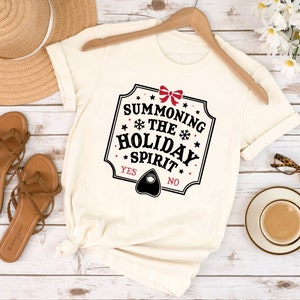 May include: Cream-colored t-shirt with the text "Summoning the Holiday Spirit" in a black, red, and white design. The shirt features a Ouija board planchette graphic and is displayed on a wooden hanger.