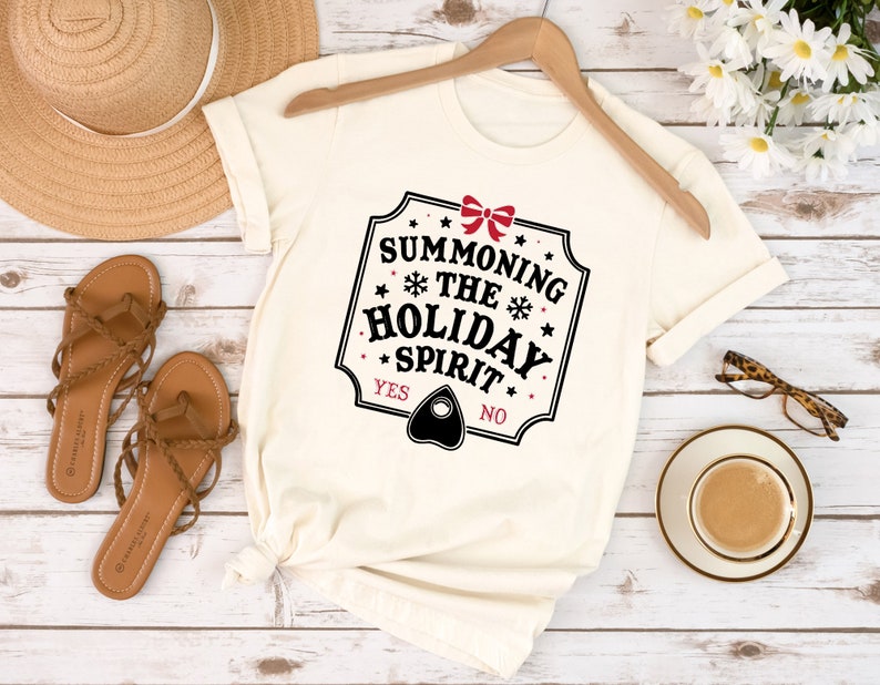 May include: Cream-colored t-shirt with the text "Summoning the Holiday Spirit" in a black, red, and white design. The shirt features a Ouija board planchette graphic and is displayed on a wooden hanger.