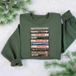 May include: A dark green sweatshirt with a stack of vintage cassette tapes printed on the front. The tapes feature artists such as Madonna, Cyndi Lauper, and Van Halen.