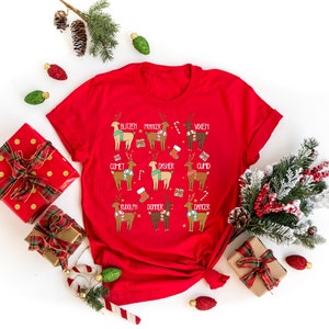 May include: A red t-shirt featuring a festive design with reindeer illustrations, each labeled with a name like "Blitzen" and "Rudolph." The reindeer are adorned with scarves and holiday accessories. The shirt is surrounded by Christmas decorations.