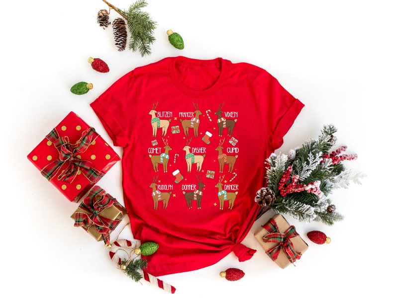 May include: A red t-shirt featuring a festive design with reindeer illustrations, each labeled with a name like "Blitzen" and "Rudolph." The reindeer are adorned with scarves and holiday accessories. The shirt is surrounded by Christmas decorations.