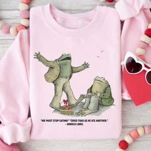 Peut inclure: Sweat-shirt rose avec un graphique de deux grenouilles du livre 'Frog and Toad are Friends' d'Arnold Lobel. Le texte sur le sweat-shirt est "We must stop eating!" "Cried Toad as he ate another." - Arnold Lobel