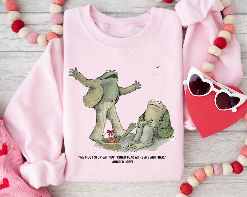 Peut inclure: Sweat-shirt rose avec un graphique de deux grenouilles du livre 'Frog and Toad are Friends' d'Arnold Lobel. Le texte sur le sweat-shirt est "We must stop eating!" "Cried Toad as he ate another." - Arnold Lobel