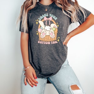 K&ouml;nnte beinhalten: Ein dunkelgraues T-Shirt mit einem Aufdruck eines wei&szlig;en Kaninchens mit einer rosa Schleife in einem Korb mit bunten Ostereiern. Der Text "Shake Your Cotton Tail" ist auf dem Shirt gedruckt.