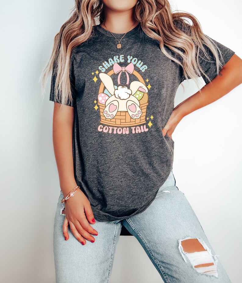 K&ouml;nnte beinhalten: Ein dunkelgraues T-Shirt mit einem Aufdruck eines wei&szlig;en Kaninchens mit einer rosa Schleife in einem Korb mit bunten Ostereiern. Der Text "Shake Your Cotton Tail" ist auf dem Shirt gedruckt.