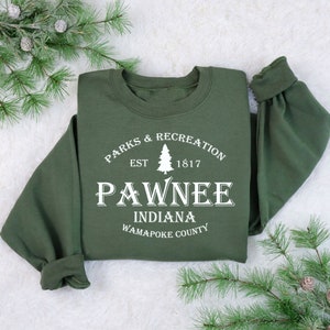Peut inclure: Un sweat-shirt vert avec un texte blanc qui dit "Parks & Recreation Est. 1817 Pawnee Indiana Wapapoke County".
