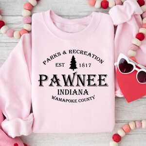 Peut inclure: Sweat-shirt rose avec un texte noir qui dit "Parks & Recreation Est 1817 Pawnee Indiana Wapapoke County". Le texte est dans une police de caract&egrave;res de style vintage et comprend un petit graphique de pin.