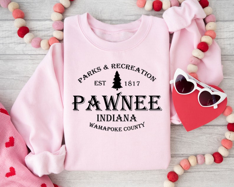 Peut inclure: Sweat-shirt rose avec un texte noir qui dit "Parks & Recreation Est 1817 Pawnee Indiana Wapapoke County". Le texte est dans une police de caract&egrave;res de style vintage et comprend un petit graphique de pin.