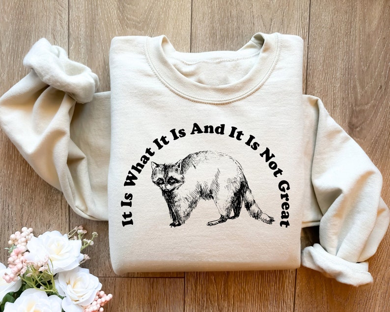 May include: Cream-colored sweatshirt featuring a black and white illustration of a raccoon. The text "It Is What It Is And It Is Not Great" is arched above the raccoon. The sweatshirt has long sleeves and a crew neck.