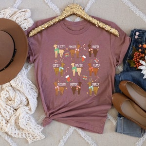 May include: A mauve-colored t-shirt featuring illustrations of reindeer with names like Blitzen, Comet, and Rudolph. The reindeer are adorned with festive accessories. The shirt is displayed with a brown hat, jeans, and flats.