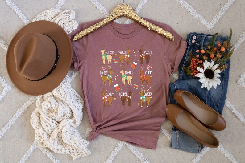 May include: A mauve-colored t-shirt featuring illustrations of reindeer with names like Blitzen, Comet, and Rudolph. The reindeer are adorned with festive accessories. The shirt is displayed with a brown hat, jeans, and flats.