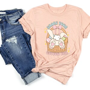 K&ouml;nnte beinhalten: Ein pinkfarbenes T-Shirt mit einem Aufdruck eines Hasen in einem Korb mit Ostereiern. Der Text auf dem Shirt lautet "Shake Your Cotton Tail".