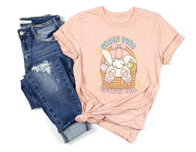 K&ouml;nnte beinhalten: Ein pinkfarbenes T-Shirt mit einem Aufdruck eines Hasen in einem Korb mit Ostereiern. Der Text auf dem Shirt lautet "Shake Your Cotton Tail".