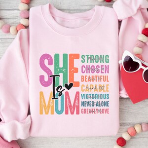 May include: Pink sweatshirt with the text "SHE IS MOM" in rainbow colours. The text is surrounded by words like "strong", "chosen", "beautiful", "capable", "victorious", "never alone", and "greatly loved".