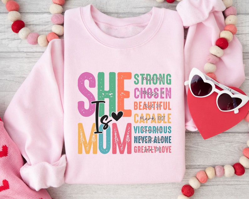 May include: Pink sweatshirt with the text "SHE IS MOM" in rainbow colours. The text is surrounded by words like "strong", "chosen", "beautiful", "capable", "victorious", "never alone", and "greatly loved".