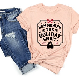 May include: A light pink t-shirt with the text "SUMMONING THE HOLIDAY SPIRIT" in a black and white design. The shirt is paired with blue denim jeans.