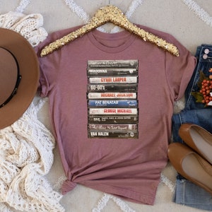 May include: A mauve t-shirt with a stack of vintage cassette tapes featuring artists like Madonna, Cyndi Lauper, and Van Halen.