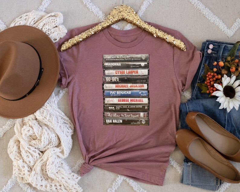 May include: A mauve t-shirt with a stack of vintage cassette tapes featuring artists like Madonna, Cyndi Lauper, and Van Halen.