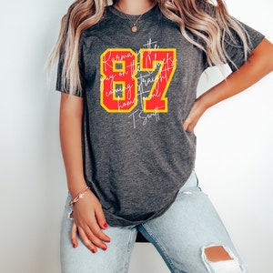 Może przedstawiać: Szary T-shirt z czerwono-ż&oacute;łtym nadrukiem, na kt&oacute;rym widnieje napis "Karma is the guy on the Chiefs coming straight home to me - T. Swift" oraz liczba 87.