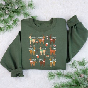 May include: An olive green sweatshirt featuring a festive design with illustrations of reindeer, each labeled with a name. The reindeer are adorned with scarves and holiday accents. The names include Blitzen, Prancer, Vixen, Comet, Dasher, Cupid, Rudolph, Donner, and Dancer.