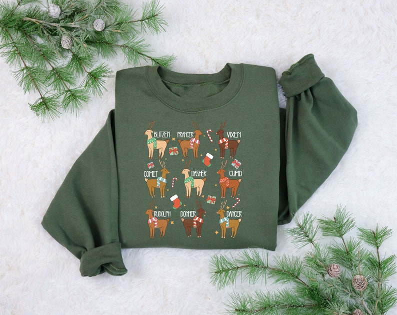 May include: An olive green sweatshirt featuring a festive design with illustrations of reindeer, each labeled with a name. The reindeer are adorned with scarves and holiday accents. The names include Blitzen, Prancer, Vixen, Comet, Dasher, Cupid, Rudolph, Donner, and Dancer.