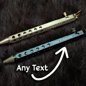 May include: Two metal pens with a series of holes along the body. One pen is gold-colored with the text "Property of Paul J". The other pen is dark blue with the text "Tony Ferguson" and the words "Any Text" with an arrow.
