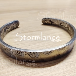 Könnte beinhalten: Ein silbernes Armband mit einem wirbelnden Muster. Das Armband ist aus Metall und hat eine glatte Oberfläche.