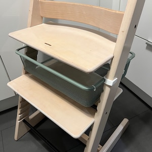 Può includere: Una sedia alta in legno bianco con un cestino in rete verde attaccato al lato. La sedia ha un vassoio rimovibile e un poggiapiedi.