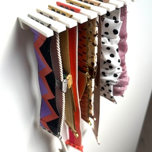 Könnte beinhalten: Ein weißer, an der Wand befestigter Organizer, der verschiedene Uhrenarmbänder zeigt. Die Bänder sind in einer Vielzahl von Farben und Mustern gehalten, darunter ein lila-oranges Zickzack-Design, eine goldene Kette und ein schwarz-weißes Polka-Dot-Muster.