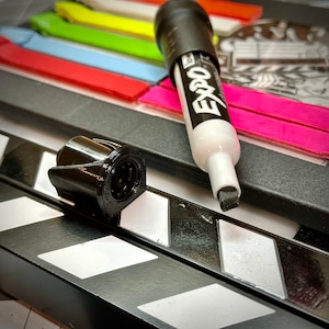 May include: A black 3D-printed object, a white and black Expo dry-erase marker, and a film clapperboard. The clapperboard has white and black diagonal stripes. Colorful sticky notes are in the background.