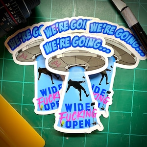 May include: A stack of stickers featuring a gray UFO with the text "WE'RE GOING..." in blue. Below, a silhouette is being beamed up with the words "WIDE FUCKING OPEN" in pink and blue. The stickers are on a green surface.