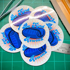 May include: A collection of round stickers featuring a blue and purple graphic of a rolled-up sleeping bag. The stickers have the text "Be Kind Rewind" in a retro font, surrounded by a sunburst design. Accessories include a ruler, tweezers, and a squeegee.
