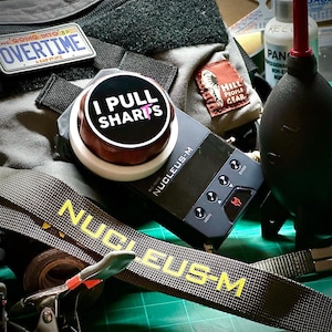 May include: An assortment of tactical gear, including a black Nucleus-M device, a strap with "NUCLEUS-M" in yellow, and a button that says "I PULL SHARPS". Other items include patches and a black air blower.