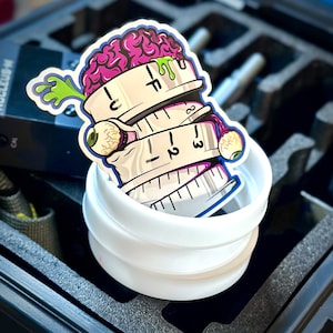 May include: A sticker featuring a cartoon brain with a green, oozing substance, atop a stack of measuring tapes. The measuring tapes have eyeballs and the numbers 1, 2, 3, and 4. The sticker is inside a white container.