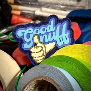 Op de afbeelding: Een blauwe, lichtblauwe en bruine patch met de tekst "Good nuff" en een duim omhoog gebaar. De patch is omgeven door rollen gekleurde tape in tinten groen, grijs en rood.