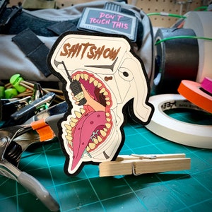 May include: A black and white patch with the text "SHIT SHOW" above a cartoon image of a mouth with a tongue and teeth. A hand holding a walkie-talkie with the words "SAVE ME" is inside the mouth. A second patch reads "DON'T TOUCH THIS".