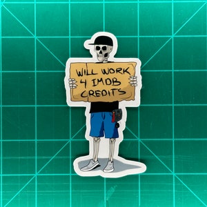 May include: A sticker depicting a cartoon skeleton in a black cap, shirt, and blue shorts, holding a sign saying "WILL WORK 4 IMDB CREDITS." The skeleton wears gray trainers and a tool belt. The sticker is on a green cutting mat.