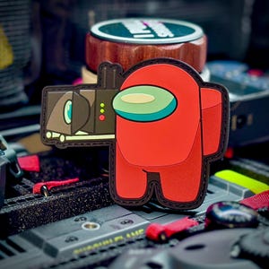 Może przedstawiać: Czerwona naszywka z postacią Among Us z mocowaniem do aparatu. Naszywka przedstawia czerwoną postać z jasnozielonym i niebieskim wizjerem. Aparat jest brązowo-czarny z zielonymi i żółtymi akcentami. Naszywka jest prawdopodobnie wykonana z wytrzymałego materiału.