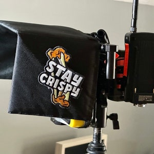 Puede incluir: Capucha negra para monitor de vídeo con el texto "STAY CRISPY" y un gráfico de comida frita. La capucha está unida a un dispositivo de vídeo negro con antenas y detalles en naranja, montado en un soporte metálico.